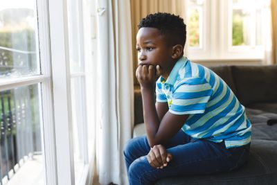 Thoughtful african american boy sitting and looking out of window in living room. spending time alone at home.